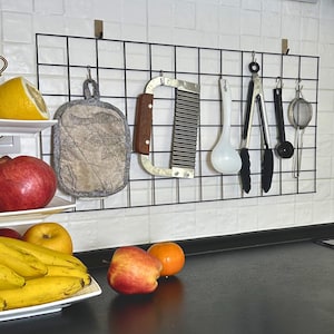 Puede incluir: Una pared de rejilla metálica negra montada en una pared de azulejos blancos con varias herramientas de cocina colgadas. Las herramientas incluyen una cuchara blanca, unas pinzas negras, una cuchara medidora negra, un colador negro y una herramienta de cocina plateada con un mango de madera.