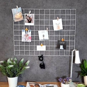 May include: A white grid memo board with photos and cards clipped to it, mounted on a grey wall. A desk with plants, a lamp, and a candle is in front of the wall. The desk is made of wood.