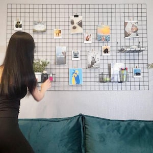 May include: A black metal grid wall organiser displaying various photos and small items clipped to it. The photos include landscapes and artwork. Two small wire baskets hold plants and other items. A person is seen near the organiser.
