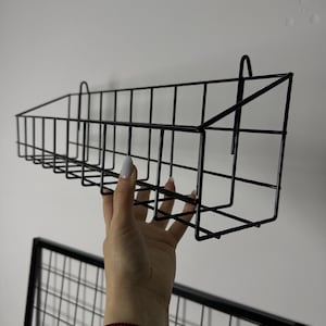 May include: A black metal wire basket with a rectangular shape, designed for wall mounting. The basket is held up by a hand. The background is a plain white wall. A second black metal wire grid is visible below the basket.