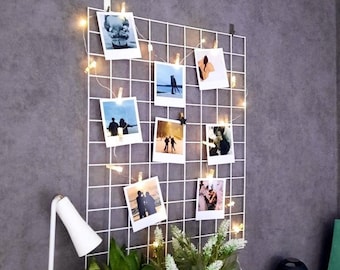 Op de afbeelding: Een wit draadgaas met lichtslingers en meerdere foto's die eraan zijn geclipte. De foto's zijn van stellen en landschappen. Het gaas is gemonteerd op een grijze muur.