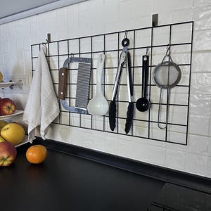 Pode incluir: Uma grade organizadora de parede em metal preto montada numa parede de azulejos brancos, contendo um pano de cozinha branco, um raspador de massa em metal com cabo de madeira, uma concha branca, pinças de cozinha pretas, uma colher medidora e um coador.