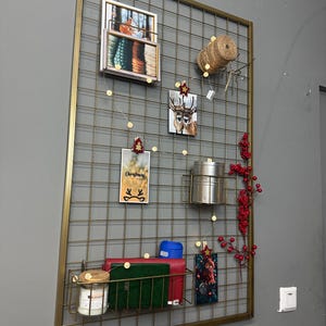 May include: A gold metal grid wall organizer with various items attached. Includes framed photos, cards, a small metal basket with a candle and books, a spool of twine, and decorative berries. Fairy lights are strung across the grid.