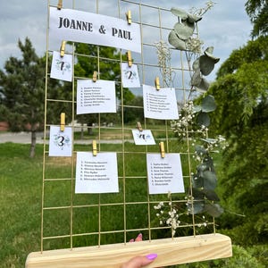 Può includere: Un tableau segnaposto a griglia metallica dorata con i nomi "JOANNE & PAUL" in alto. Piccole carte bianche con i nomi degli ospiti sono attaccate alla griglia. Il tableau è decorato con verde e poggia su una base di legno.