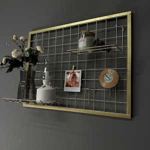 May include: A gold metal grid wall organiser with two shelves. The shelves hold a candle, a photo, a small clock, and a vase of flowers. A white lamp is also on the shelf. The organiser is mounted on a grey wall.