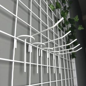 Op de afbeelding: Witte metalen roosterwand met verschillende gebogen haken, ideaal voor het organiseren of weergeven van items. Het rooster is gemonteerd op een grijze muur, met groene bladranken op de achtergrond. De haken zijn ontworpen om verschillende objecten vast te houden.