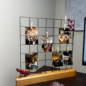 Può includere: Un espositore per foto a griglia metallica nera con base in legno chiaro. La griglia presenta cinque fotografie rettangolari verticali fissate con piccole mollette di legno. Rametti decorativi di bacche rosse sono fissati al telaio.