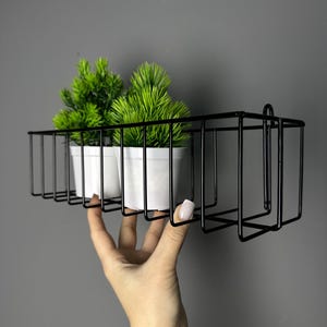 Op de afbeelding: Een zwart metalen draadplank met twee kleine witte potten met groene planten. De plank heeft een rechthoekig ontwerp met een vierkant gedeelte aan de rechterkant. De plank wordt door een hand tegen een grijze achtergrond gehouden.