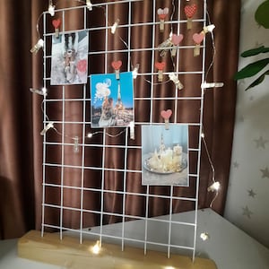 May include: A white wire grid photo display with a wooden base, decorated with mini pegs and heart-shaped clips. Photos are attached, and a string of fairy lights provides a warm glow. The display is set against a brown curtain.