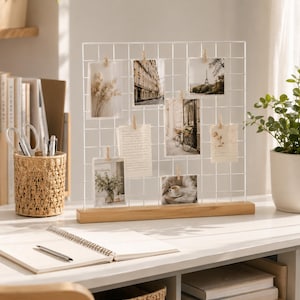 Pode incluir: Um expositor de fotos em grade de arame branco com uma base retangular de madeira clara. A grade possui vários pequenos clipes de madeira que prendem diversas fotografias e notas, incluindo imagens de uma rua, a Torre Eiffel e flores.