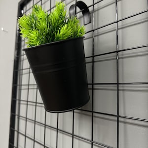 May include: A black metal hanging planter with a bright green plant. The planter is suspended from a black grid wall. The plant has a full, textured appearance, contrasting with the smooth, dark surface of the planter.