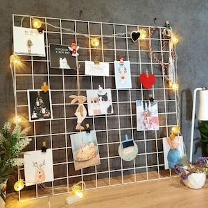 Op de afbeelding: Een witte metalen roosterwand met lichtslingers en verschillende items die eraan vastgemaakt zijn, waaronder foto's, kaarten en een houten konijnenfiguur. De roosterwand staat op een houten oppervlak.