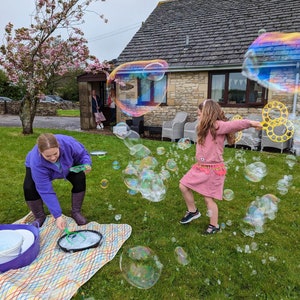 May include: A young girl in a pink dress blows bubbles with a yellow bubble wand in a grassy garden. A woman in a purple jacket is making more bubbles with a bucket and wand.