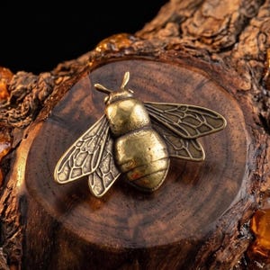 May include: A detailed, close-up shot of a brass bee figurine resting on a dark wood surface. The bee has intricate wing and body details. Amber-colored resin droplets are scattered around the wood, adding a natural element to the composition.