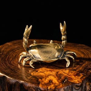 May include: A small, detailed brass crab figurine with raised claws. The crab is positioned on a dark, textured wooden surface, set against a black background. The crab's legs and claws are clearly defined, showcasing the craftsmanship.