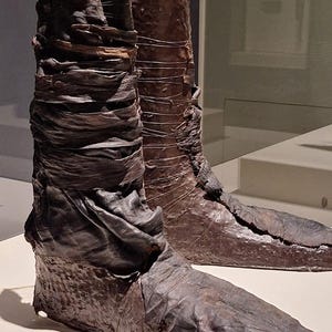 May include: A pair of brown leather boots with a unique design. The boots are made of multiple layers of leather, which have been stitched together. The boots are on display in a museum.