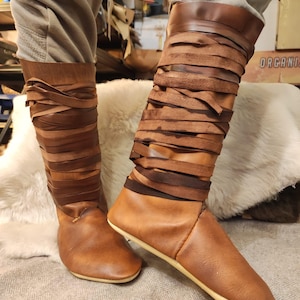 May include: A pair of brown leather boots with a unique design. The boots are laced up the calf with brown leather straps. The boots have a pointed toe and a flat sole.