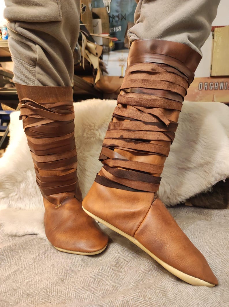 May include: A pair of brown leather boots with a unique design. The boots are laced up the calf with brown leather straps. The boots have a pointed toe and a flat sole.