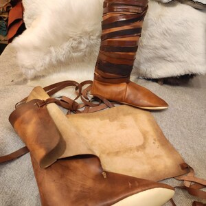 May include: A pair of brown leather boots with laces. The boots are made of thick leather and have a pointed toe. The boots are sitting on a white fur rug.