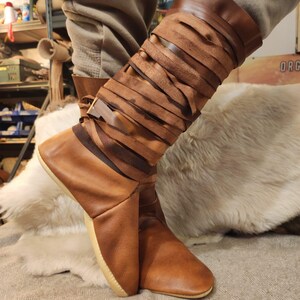 May include: Brown leather boots with a unique design featuring multiple straps wrapped around the calf. The boots have a pointed toe and a light brown sole.