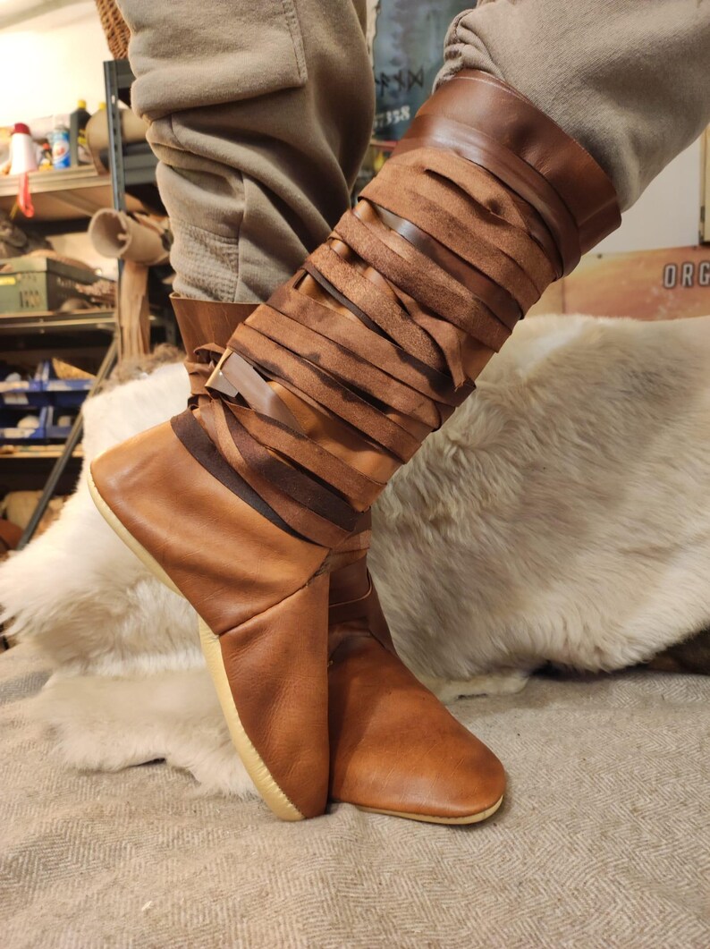May include: Brown leather boots with a unique design featuring multiple straps wrapped around the calf. The boots have a pointed toe and a light brown sole.