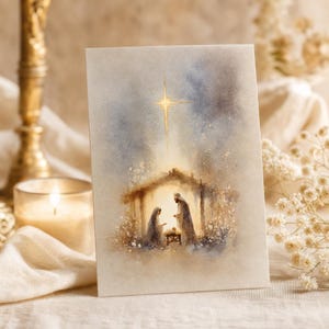 May include: A rectangular greeting card featuring a watercolour illustration of a nativity scene with a bright star above a stable, depicting two figures standing over a manger in soft shades of blue, gold, and cream on a textured paper surface.
