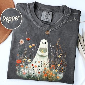 May include: Dark grey Comfort Colors t-shirt featuring a whimsical ghost holding a book with a crescent moon and stars. The ghost is surrounded by colorful wildflowers. The word "Pepper" is in a circle on the upper left.