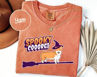 May include: Orange Comfort Colors t-shirt with a graphic of a corgi wearing a witch hat and riding a broom, with the text "Spooky Cooorgi". A gold necklace and a brown hat are also visible. The shirt is a size small and the color is "Yam".