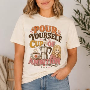 May include: A cream-colored t-shirt featuring a graphic print with the text "POUR YOURSELF A CUP OF AMBITION" and "9 TO 5". The design includes a coffee cup with a flower, a moka pot, and an illustration of a blonde woman in a pink shirt.