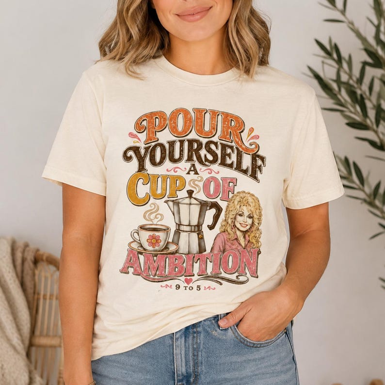 May include: A cream-colored t-shirt featuring a graphic print with the text "POUR YOURSELF A CUP OF AMBITION" and "9 TO 5". The design includes a coffee cup with a flower, a moka pot, and an illustration of a blonde woman in a pink shirt.