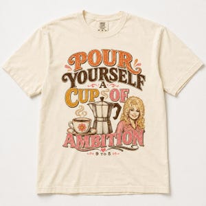 May include: A cream-colored short-sleeve t-shirt featuring a graphic print with the text "POUR YOURSELF A CUP OF AMBITION" and "9 TO 5". The design includes a coffee cup, a moka pot, and an illustration of a blonde person in a pink shirt.