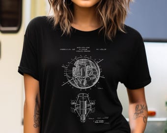 Può includere: Una t-shirt nera con un'illustrazione tecnica di brevetto in bianco di una struttura satellitare. La grafica include disegni al tratto dettagliati dei componenti del satellite, insieme al testo stampato United States Patent, R.C. Baumann, Satellite Structure, Pat. 2,921,258 e Patented Jan. 19, 1960.