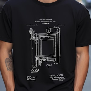 May include: A black t-shirt featuring a white illustration of a patent drawing for an elevator. The drawing is labelled "ELEVATOR" and includes the text "PATENTED MAY 25, 1880" and "PAT. 228,107". The drawing is signed "Charles R. Otis" and "By his attorney, Charles E. Otis".