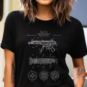 May include: A black t-shirt featuring a white technical patent illustration of a Heckler & Koch MP5 firearm. The graphic includes detailed schematics, numbered parts, and technical text, including the patent date of February 26, 1985.