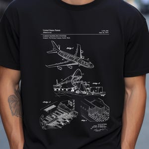 Puede incluir: Camiseta negra con un esquema blanco de un sistema de manipulación de carga de aviones. El diseño incluye diagramas del avión y sus mecanismos de carga de carga, con información de patente. La camiseta tiene cuello redondo.