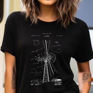 Puede incluir: Una camiseta negra de manga corta con un estampado gráfico blanco que muestra una ilustración técnica de patente para una turbina eólica. El diseño incluye dibujos lineales detallados de la estructura, componentes y texto.