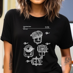 Op de afbeelding: Een zwart T-shirt met korte mouwen en een witte grafische print van een Amerikaans patent voor een schijnwerper. Het ontwerp bevat technische lijntekeningen met de labels Fig. 1, Fig. 2, Fig. 3 en Fig. 4, samen met patenttekstdetails.