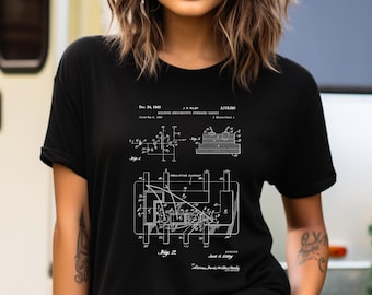 Può includere: Una t-shirt nera a maniche corte con una stampa grafica bianca di un'illustrazione di brevetto del 1963 per un circuito integrato a semiconduttore in miniatura. Il design include schemi tecnici, etichette e il testo Dec. 24, 1963, J.S. Kilby, 3,115,561.