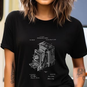 May include: A black short-sleeved t-shirt featuring a white graphic print of a vintage photographic camera patent illustration. The design includes technical line drawings of the camera and text reading T.H. BLAIR PHOTOGRAPHIC CAMERA.