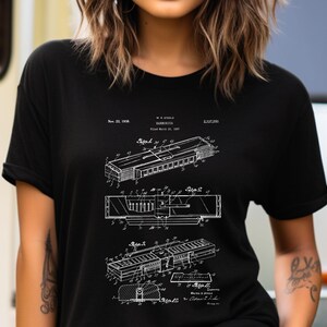 May include: A black t-shirt featuring a white technical patent illustration of a harmonica. The graphic includes detailed line drawings labelled Fig. 1 through Fig. 4, along with the text Nov. 22, 1938, 2,137,251, and Harmonica, Filed March 10, 1937.
