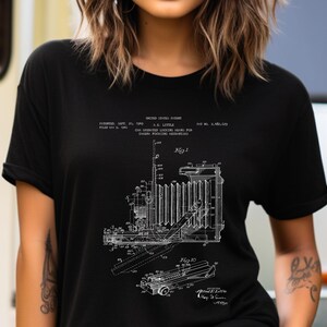 Può includere: Una t-shirt nera a maniche corte con una stampa grafica bianca di un brevetto statunitense per un meccanismo di messa a fuoco di una fotocamera. Il design include disegni tecnici a linee etichettati Fig 1 e Fig 10, insieme al testo del brevetto.