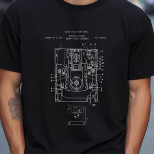 May include: A black t-shirt featuring a white technical patent illustration of a floppy disk assembly. The graphic includes the text United States Patent Office, Takanasa Kitano, Floppy Disk Assembly, Patented May 13, 1997, and Pat. 5,629,819.