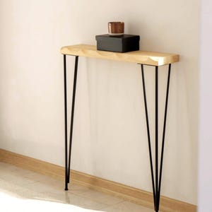 May include: A light wood console table with black hairpin legs. The table has a natural wood finish and a slightly curved front edge. A black box and a small brown cup sit on top of the table. The table is against a white wall.