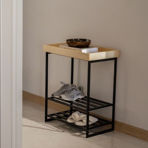 Pu&ograve; includere: Un porta scarpe in metallo nero con due ripiani e un piano in legno. Il ripiano superiore ha una piccola ciotola marrone e un libro con la parola "Federal" visibile. Il porta scarpe &egrave; su un tappeto bianco davanti a un muro bianco.