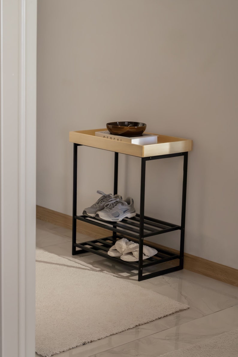 Pu&ograve; includere: Un porta scarpe in metallo nero con due ripiani e un piano in legno. Il ripiano superiore ha una piccola ciotola marrone e un libro con la parola "Federal" visibile. Il porta scarpe &egrave; su un tappeto bianco davanti a un muro bianco.