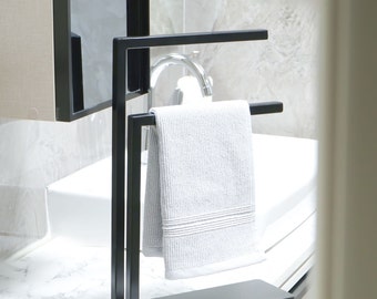 May include: A black metal towel rack with two bars, a white towel hanging on the bottom bar, and a white porcelain sink in the background.