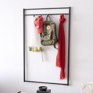 Può includere: Un portabiti da parete in metallo nero con un berretto da baseball rosso, uno zaino verde e una sciarpa rossa appesi. Una borsa a rete bianca con frutta verde e gialla è appesa a sinistra. Una ciotola e una scatola nere sotto.