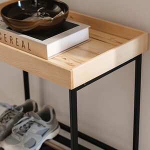 Pu&ograve; includere: Un porta scarpe in metallo nero con un ripiano superiore in legno. Sul ripiano superiore c'&egrave; una ciotola marrone e un libro intitolato "Cereal".