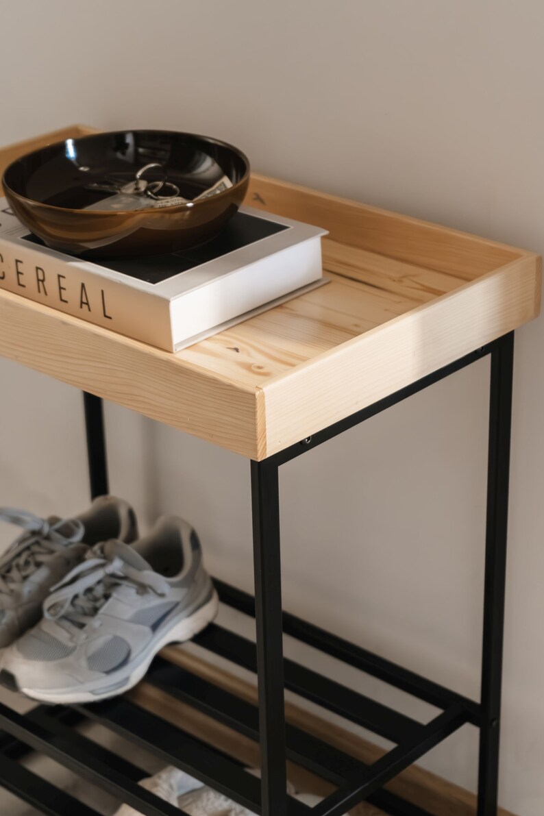 Pu&ograve; includere: Un porta scarpe in metallo nero con un ripiano superiore in legno. Sul ripiano superiore c'&egrave; una ciotola marrone e un libro intitolato "Cereal".