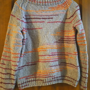 May include: A gray knit sweater with horizontal stripes in shades of orange, red, and burgundy. The sweater has a crew neck and long sleeves. The stripes are evenly spaced across the chest and sleeves, creating a textured look. The sweater is hanging on a clear plastic hanger.